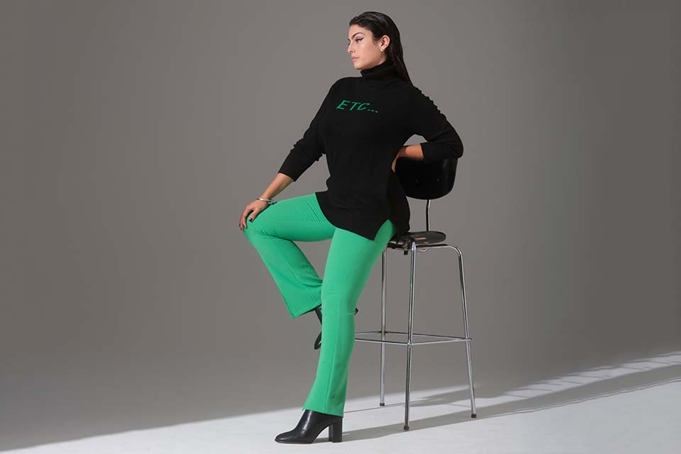 The model sits on a bar stool and wears green trousers and a black jumper with green lettering