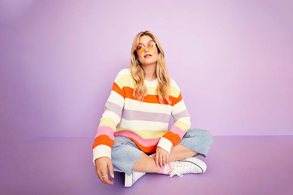 A blonde model sits on the floor wearing a striped jumper and colourful sunglasses