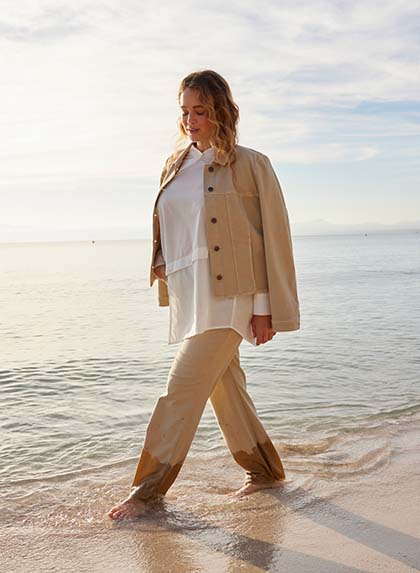 A woman walking on a beach wearing a beige two-piece and a white blouse