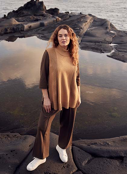 The model poses in a brown outfit in front of a body of water


