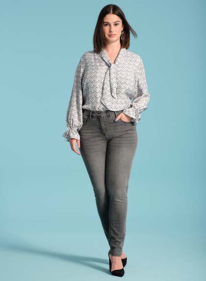 The model poses in a blouse and gray jeans in front of a blue wall 