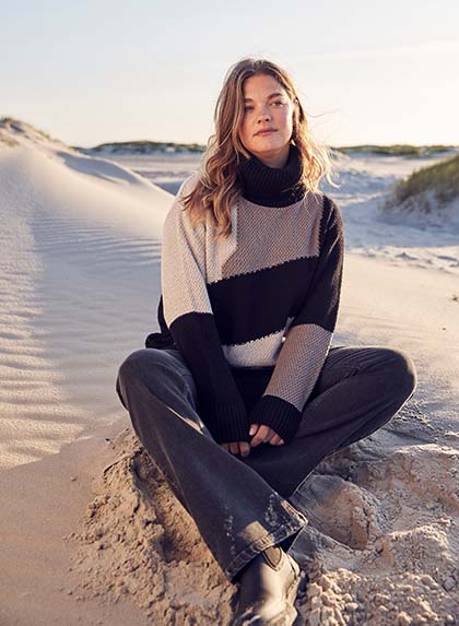 The model is sitting on the beach wearing a turtleneck sweater 


