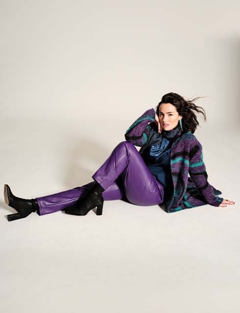 The model poses on the ground wearing purple trousers and a colourful cardigan. 

