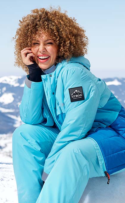 The model sits on the mountain in a blue ski jacket